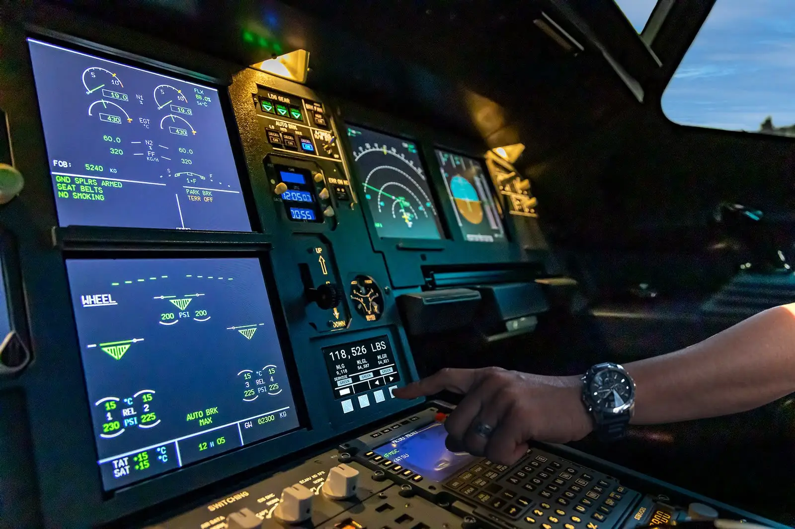 A pilot's hand pressing a button on the ES3 SHMS central control unit display in an airliner cockpit, showing gross weight and gear loads